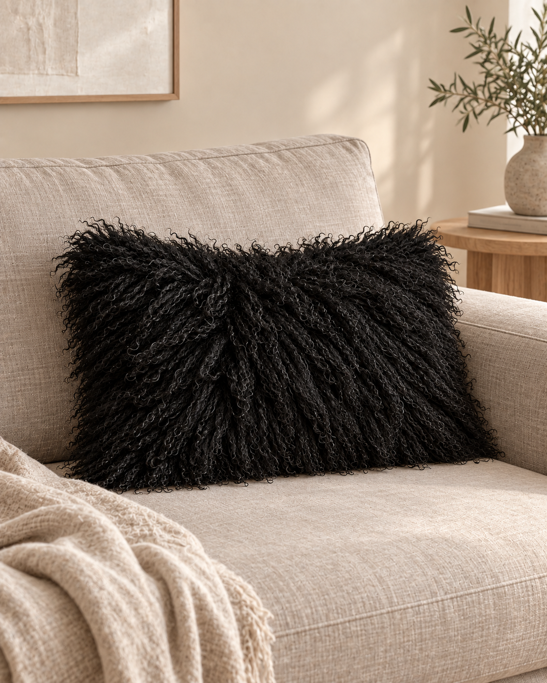 Black rectangular Mongolian sheepskin cushion on a beige sofa beside a knitted throw